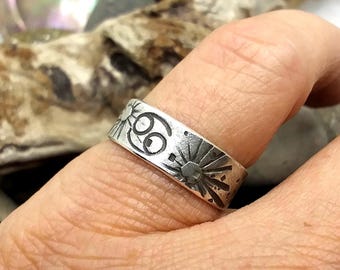 May include: A silver-toned metal ring featuring a flat band design with engraved details. The surface displays a central Cancer zodiac symbol flanked by two sunburst motifs, all rendered in a dark, oxidized finish against the silver metal.