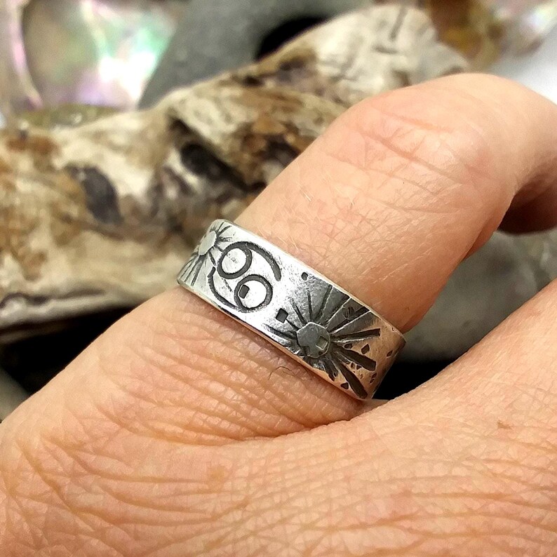 May include: A silver-toned metal ring featuring a flat band design with engraved details. The surface displays a central Cancer zodiac symbol flanked by two sunburst motifs, all rendered in a dark, oxidized finish against the silver metal.