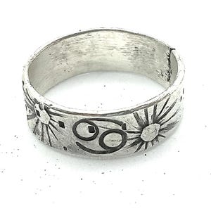 May include: A silver-toned band ring featuring a textured, brushed finish. The exterior surface is engraved with a repeating pattern of sunburst motifs and the Cancer zodiac symbol, rendered in dark, recessed lines against the metal.