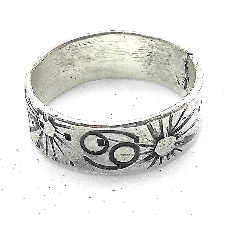 May include: A silver-toned band ring featuring a textured, brushed finish. The exterior surface is engraved with a repeating pattern of sunburst motifs and the Cancer zodiac symbol, rendered in dark, recessed lines against the metal.