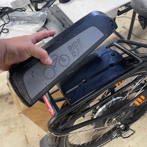 May include: A black electric bike battery with the text "RIDE RAD" and a bicycle graphic is being held near the bike frame. The bike frame is black with orange accents. The bike is partially assembled.