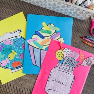Op de afbeelding: Drie kleurrijke papieren knutselwerken met een thema van eten en keukengerei. De eerste knutsel is een geel papier met een witte omtrek van een viskom gevuld met kleurrijke vissen. De tweede knutsel is een blauw papier met een witte omtrek van een cupcake met kleurrijke sprinkels. De derde knutsel is een roze papier met een witte omtrek van een pot gevuld met kleurrijk keukengerei. De pot heeft het woord "UTENSILS" erop geschreven.
