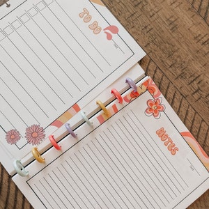 May include: A white planner with a to-do list and notes section. The planner has a colorful floral design and a date section. The planner is bound with colorful rings.