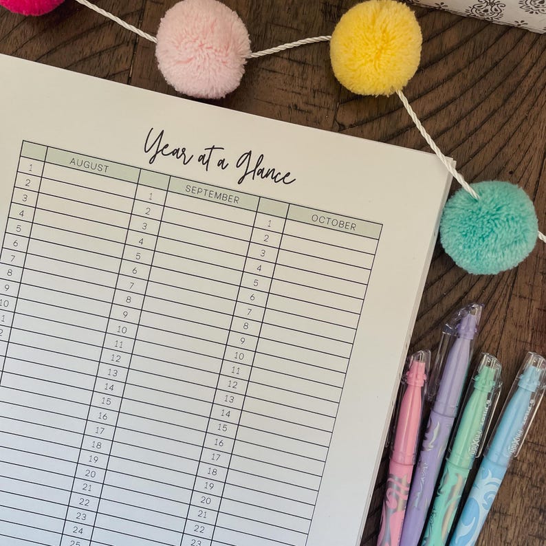 May include: A white planner with a green header that says "Year at a Glance". The planner has columns for August, September, and October, with numbered days of the month. The planner is on a wooden surface with a string of colorful pom-poms.