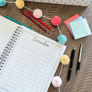 May include: A white spiral bound notebook with the word "Curriculum" written in black cursive. The notebook has columns for subject, book title, grade, publisher, level, year, and semester. The notebook is surrounded by colorful pom-poms, pencils, and pens.