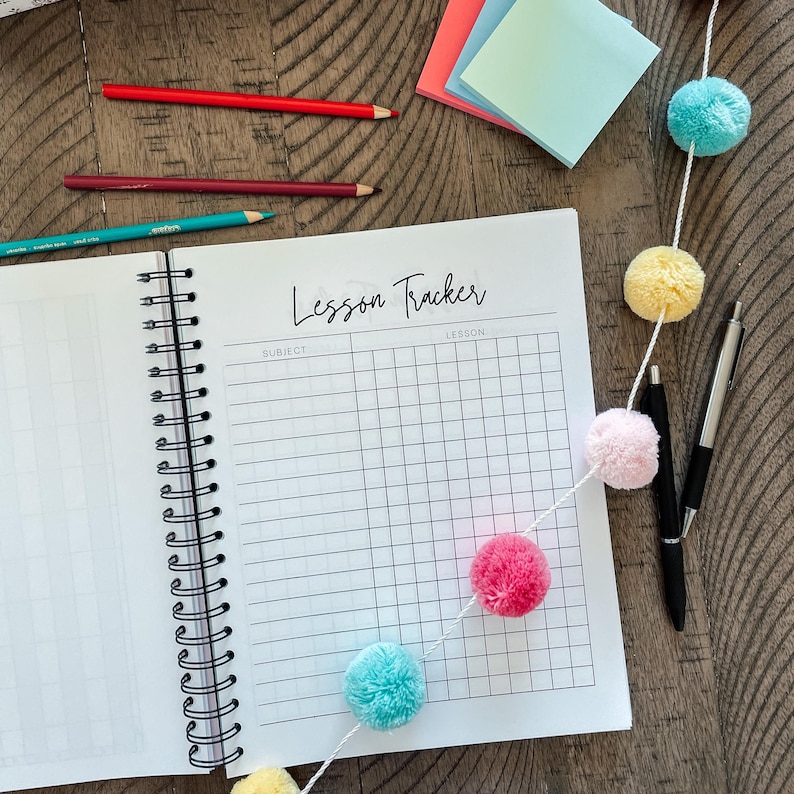 May include: A white spiral bound notebook with the words "Lesson Tracker" written in black cursive. The notebook is open to a page with a grid for tracking subjects and lessons. A string of colorful pom-poms decorates the bottom of the notebook.