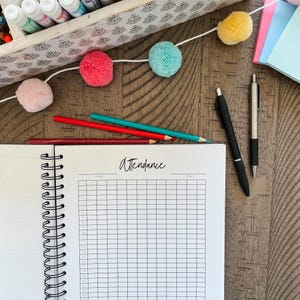 May include: A white spiral bound notebook with the word "attendance" written on the cover. The notebook is open to a blank page with a grid pattern. There are colored pencils, pens, and pom-poms on a wooden table.
