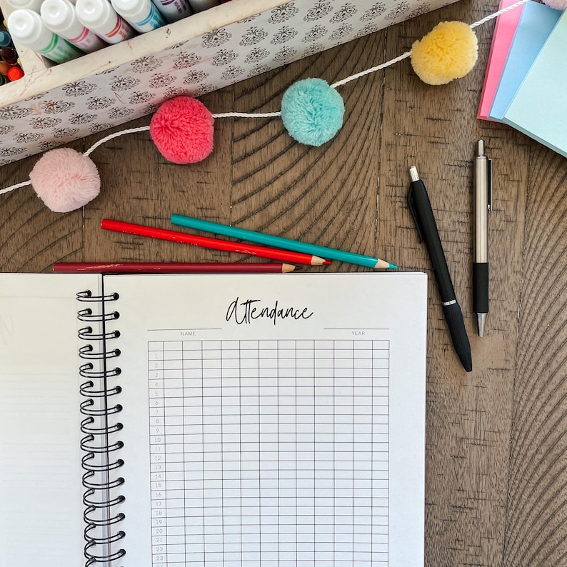 May include: A white spiral bound notebook with the word "attendance" written on the cover. The notebook is open to a blank page with a grid pattern. There are colored pencils, pens, and pom-poms on a wooden table.