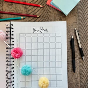 May include: A white spiral bound notebook with the words "Our Year" written in black cursive at the top. The notebook has a grid of squares for each day of the year. A string of colorful pom-poms is draped across the notebook.