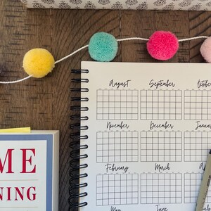 May include: A white planner with black gridded monthly calendars for August through July. The planner has a black spiral binding and is lying on a wooden surface. A string of colorful pom-poms is draped across the top of the planner.