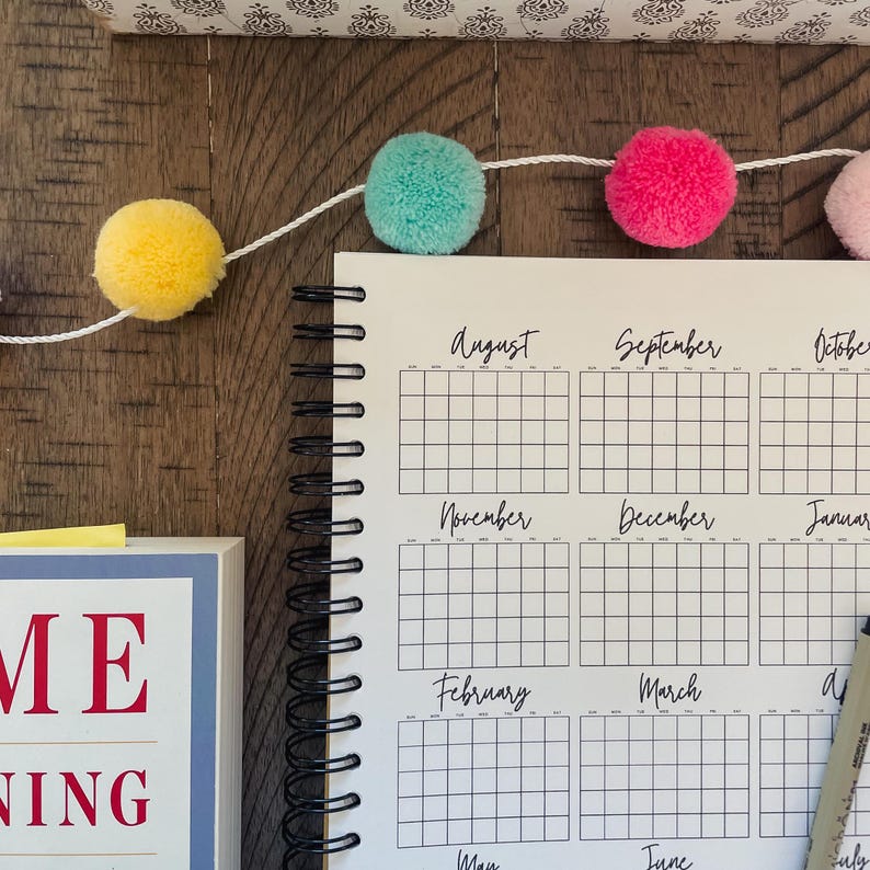 May include: A white planner with black gridded monthly calendars for August through July. The planner has a black spiral binding and is lying on a wooden surface. A string of colorful pom-poms is draped across the top of the planner.