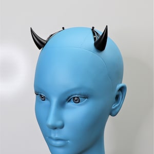 May include: Black devil horns attached to a black headband. The horns are curved and pointed, and the headband is designed to sit on the head. The horns are displayed on a blue mannequin head.