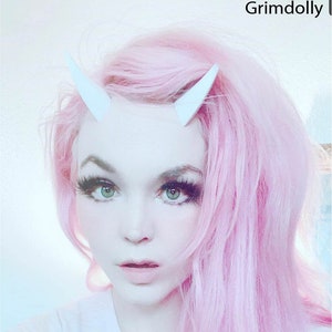 May include: A person with pink hair wearing white devil horns. The person is looking at the camera and has dark eye makeup.