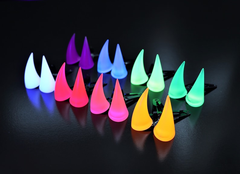 May include: A collection of colorful, glowing horn-shaped hair clips. The horns are arranged in rows, displaying a spectrum of colors including white, pink, orange, yellow, green, blue, and purple. Each clip has a black base.