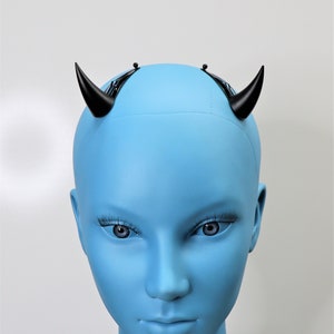 May include: A pair of black devil horns attached to a black headband, displayed on a blue mannequin head. The horns are curved and pointed, and the mannequin has realistic facial features.