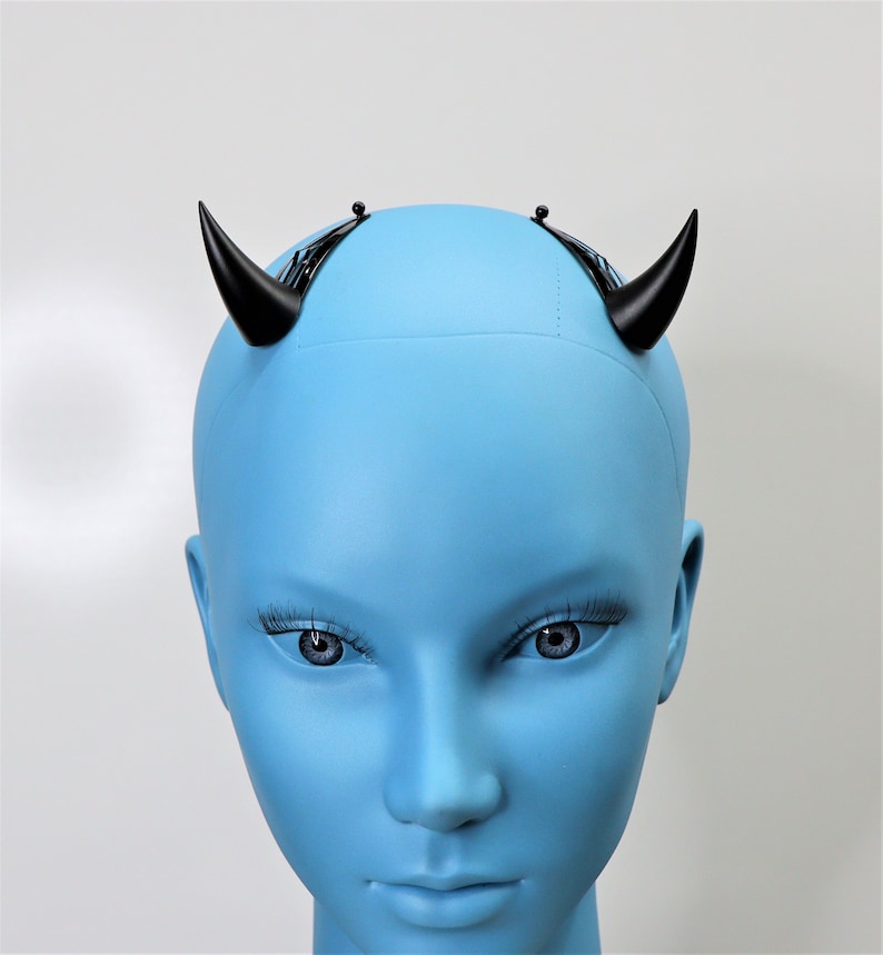 May include: A pair of black devil horns attached to a black headband, displayed on a blue mannequin head. The horns are curved and pointed, and the mannequin has realistic facial features.