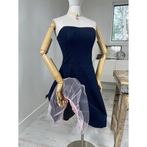 Puede incluir: Un vestido azul marino sin tirantes con falda acampanada se exhibe en un maniquí. Una tela rosa transparente está drapeada sobre el brazo del maniquí. El maniquí lleva una parte superior blanca y una gargantilla color crema con un pequeño colgante.