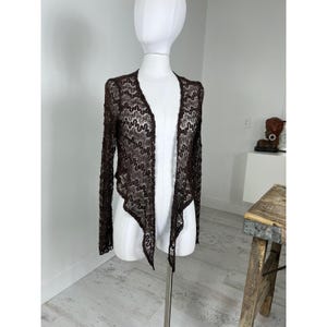 May include: A dark brown long-sleeve cardigan made of sheer lace fabric. The garment features an open front design with a wavy, vertical pattern throughout the material. The hemline is asymmetrical, with longer points at the front and long sleeves.