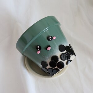 以下が含まれることがあります： かわいい漫画の顔と底に黒いタピオカパールが付いた緑色のセラミックプランター。