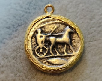 May include: A gold-toned pendant with a detailed image of a horse-drawn chariot. The pendant is oval-shaped and has a textured surface.
