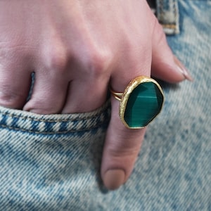 May include: A gold ring with a large, faceted, green gemstone. The ring is on a finger, and the hand is resting on a denim pocket.