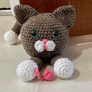 May include: A brown crocheted cat toy with white paws and a pink nose. The cat is holding a pink ball of yarn.