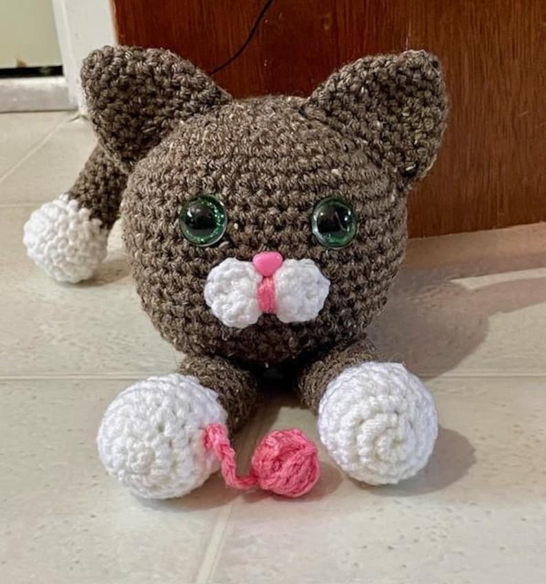May include: A brown crocheted cat toy with white paws and a pink nose. The cat is holding a pink ball of yarn.