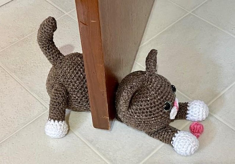 May include: A brown crocheted cat doorstop with white paws and pink accents. The cat is lying on its side with its head resting on the floor.