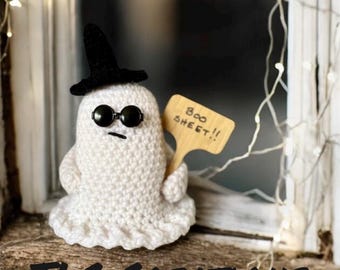 Puede incluir: Un fantasma de crochet con un sombrero de bruja negro y gafas de sol, sosteniendo un cartel de madera que dice "Boo Sheet!!".