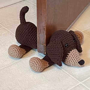 May include: A brown and tan crocheted dachshund dog doorstop. The dog is lying on its side with its head resting on the floor. The dog's tail is curled up behind it.