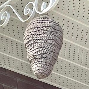 Op de afbeelding: Een handgemaakt, lichtbruin gehaakt wespennest dat aan een witte decoratieve metalen beugel hangt. Het nest is kegelvormig met horizontale rijen haaksteken. De achtergrond omvat een wit plafond en een bakstenen muur.
