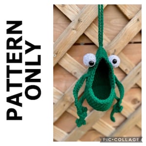 Può includere: Mangiatoia per uccelli a forma di rana in crochet verde con occhietti bianchi sporgenti appesa a un reticolo di legno.
