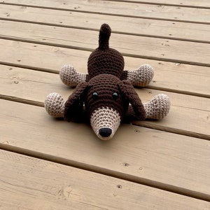May include: A brown and beige crocheted dachshund dog toy lying on a wooden surface.
