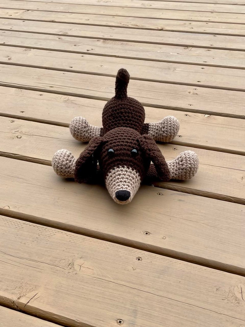 May include: A brown and beige crocheted dachshund dog toy lying on a wooden surface.