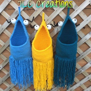 May include: Three handmade crochet wall pockets in blue, yellow, and teal, each with a fringe bottom. The pockets are suspended from a wooden lattice, with whimsical eye and antenna details. The text "TLC creations" is visible.