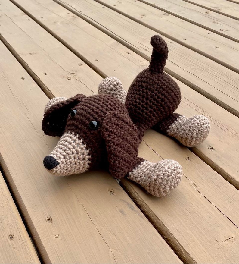 May include: A brown and tan crocheted dog toy with black eyes. The toy is lying on a wooden surface.