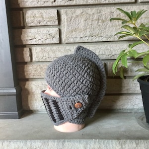 May include: Gray crocheted knight helmet hat with a black and gray striped face mask. The hat has a button detail on the side.