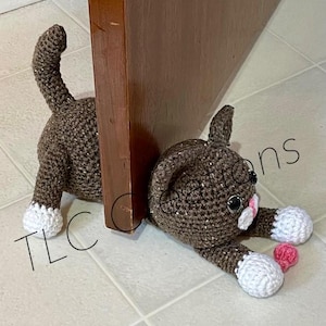 Op de afbeelding: Een bruine gehaakte kattendeurstop met witte poten en roze accenten. De kat ligt met zijn kop op de vloer. De kat is geplaatst tegen een houten deur.