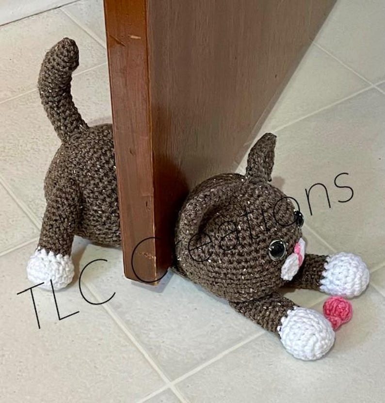 May include: A brown crocheted cat doorstop with white paws and pink accents. The cat is lying down with its head resting on the floor. The cat is positioned against a wooden door.