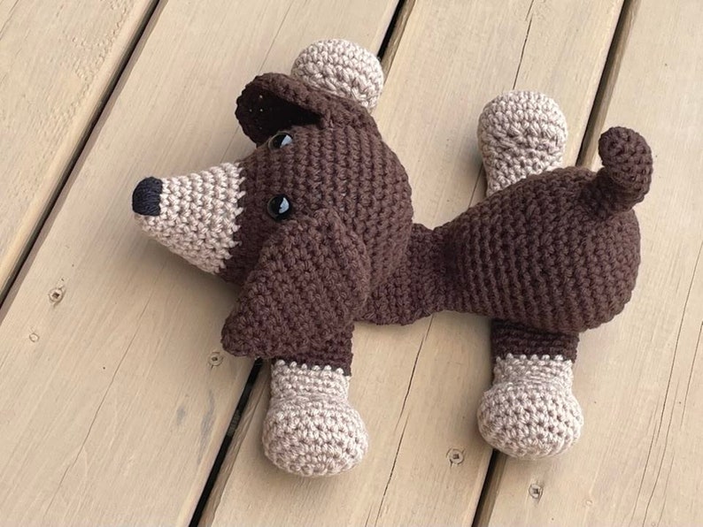 May include: A brown crocheted dog toy with beige accents. The dog is lying on its side with its head turned to the side.