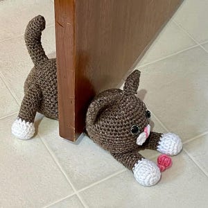 May include: A brown crocheted cat doorstop with white paws and pink nose. The cat is lying down with its head resting on the floor. The cat is holding a pink ball in its paw.