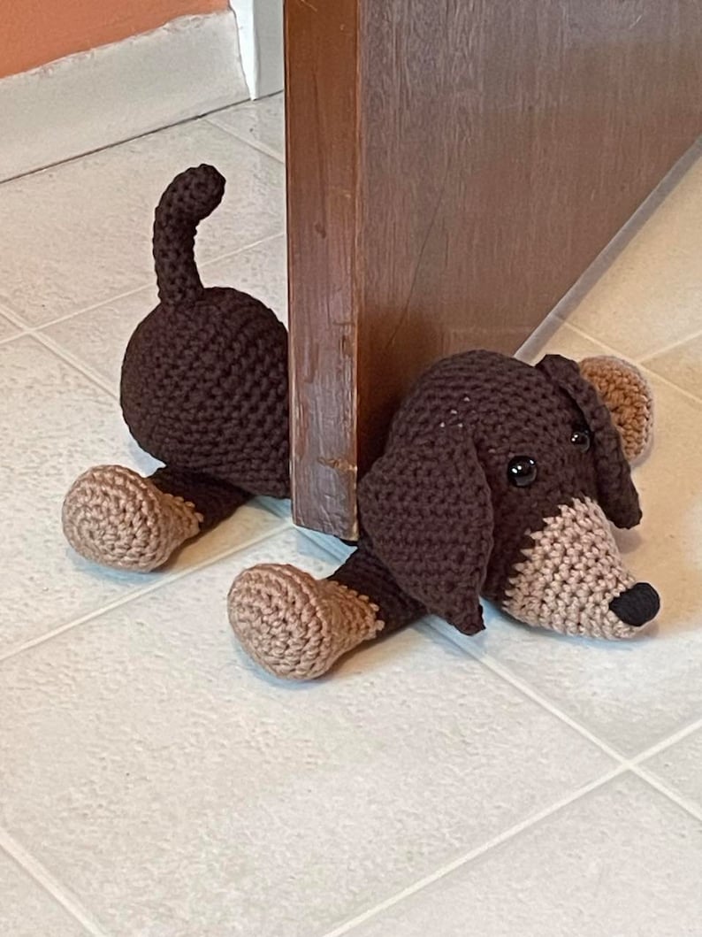 May include: A brown and tan crocheted dachshund dog doorstop. The dog is lying on its side with its head resting on the floor. The dog's tail is curled up behind it.