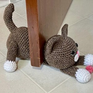 May include: A brown crocheted cat doorstop with white accents and pink details. The cat is lying down with its head resting on the floor and its tail curled up behind it.
