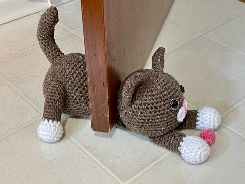 May include: A brown crocheted cat doorstop with white accents and pink details. The cat is lying down with its head resting on the floor and its tail curled up behind it.