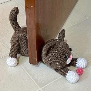 May include: A brown crocheted cat doorstop with white paws and pink accents. The cat is lying on its side with its head resting on the floor. The cat is holding a pink heart in its paw.