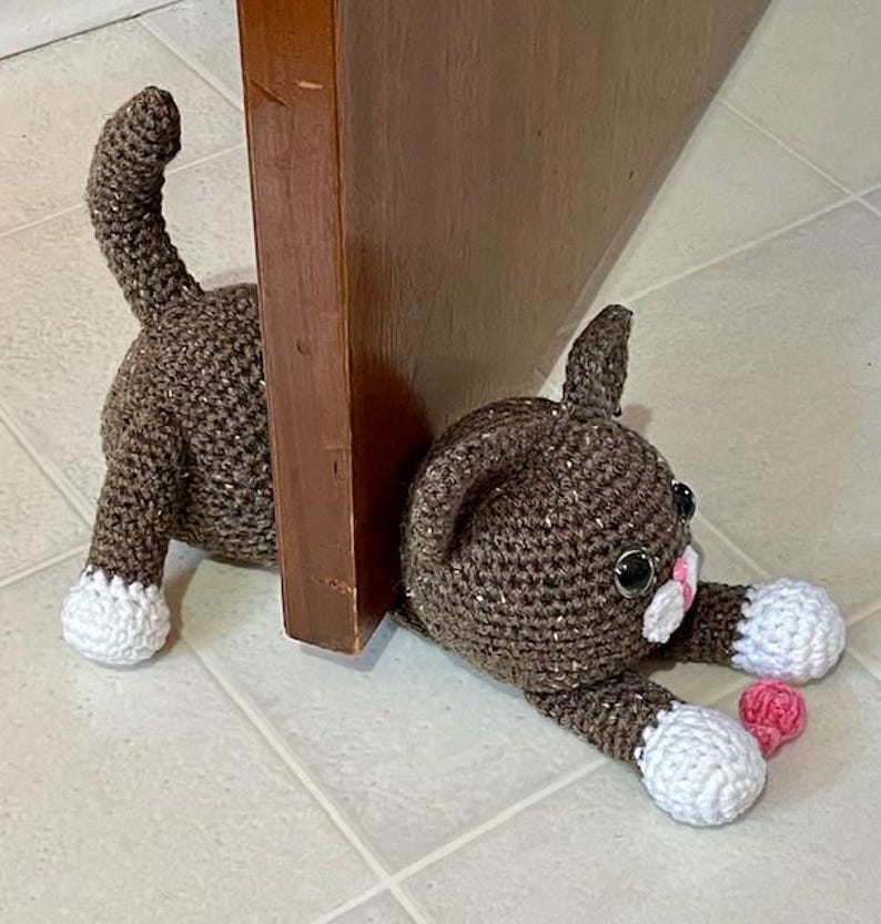 May include: A brown crocheted cat doorstop with white paws and pink accents. The cat is lying on its side with its head resting on the floor. The cat is holding a pink heart in its paw.