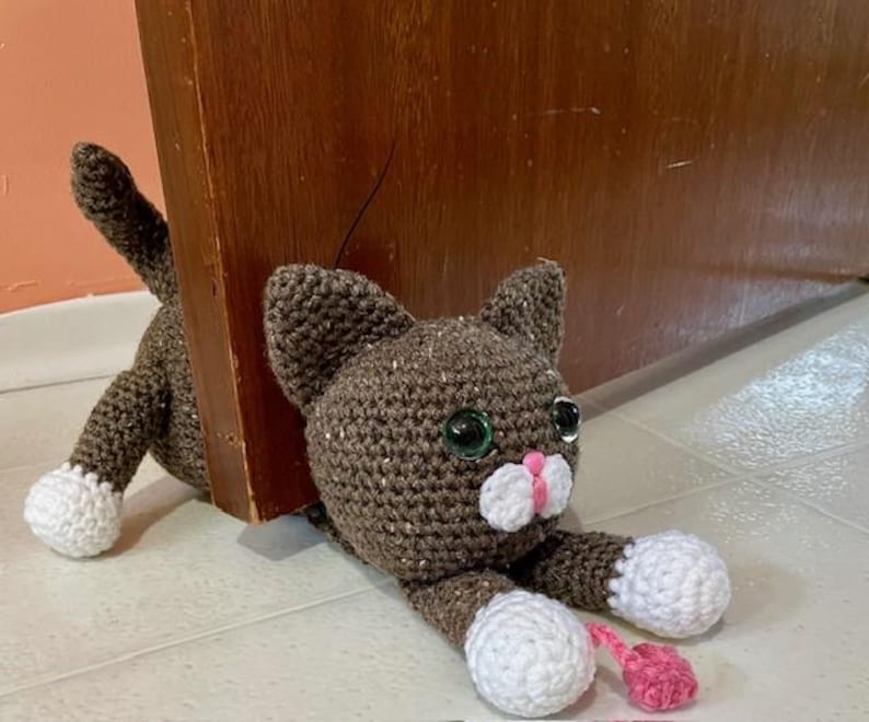 May include: A brown crocheted cat doorstop with white paws and a pink nose. The cat is holding a pink mouse in its mouth.