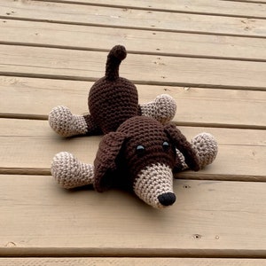 May include: A brown and beige crocheted dog toy with black eyes. The dog is lying on its belly with its legs outstretched.