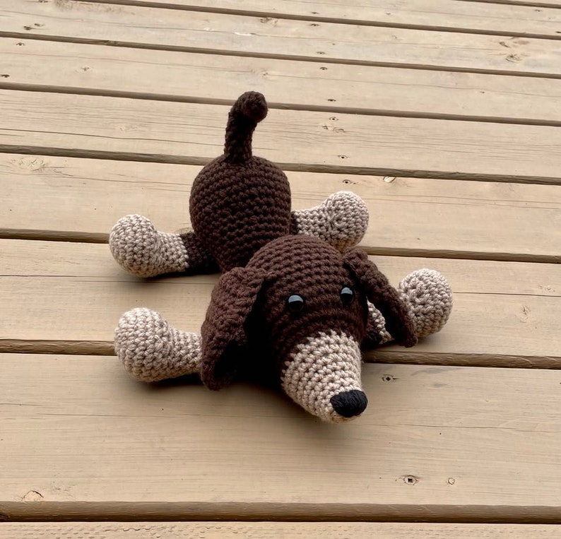 May include: A brown and beige crocheted dog toy with black eyes. The dog is lying on its belly with its legs outstretched.