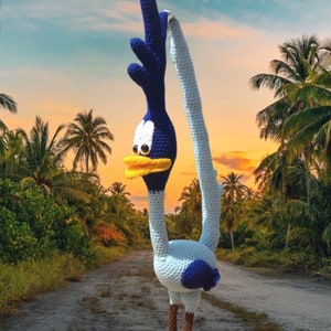 May include: A crocheted Road Runner toy with blue and white feathers, a yellow beak, and brown legs. The toy is standing on a dirt road with palm trees in the background.